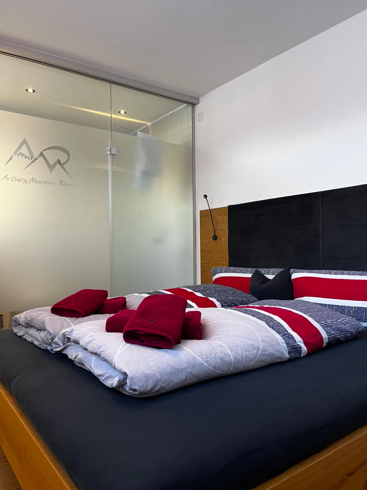 Doppelbett im Obergeschoss mit roten Kissen, dahinter das Bad hinter Glas mit dem Logo